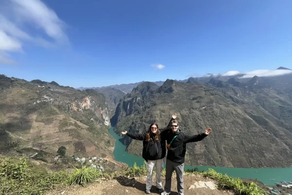 Two happy Loop Trails Tours customers posing with wide smiles and open arms on the legendary Ma Pi Leng Pass, with the breathtaking view of the Nho Que River and the Tu San Canyon – the deepest canyon in Southeast Asia – behind them. A highlight of the Ha Giang loop motorbike tour experience.